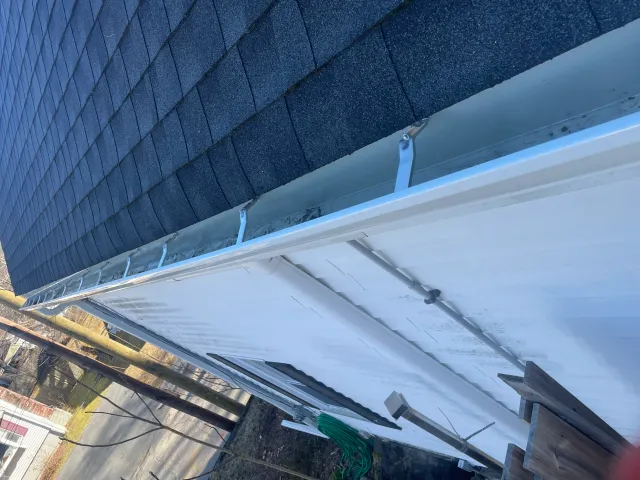 A close-up view of a house roof with asphalt shingles, white gutters, and downspouts, next to a utility pole and a road.