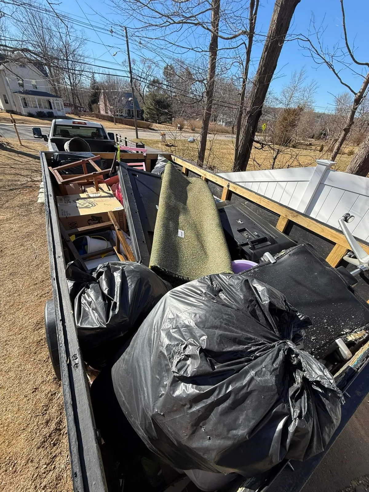 Trailer filled with debris: black trash bags, rug, wood, and other items in a sunny outdoor setting.