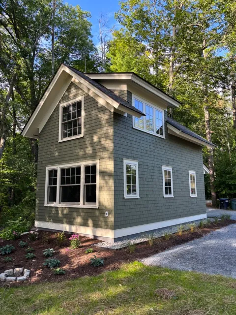 A two-story house with gray siding and white trim, surrounded by trees and a landscaped yard with flowers and mulch.