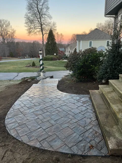 Brick walkway and stone steps leading to a suburban front yard at sunset, with shrubs and a striped pole nearby
