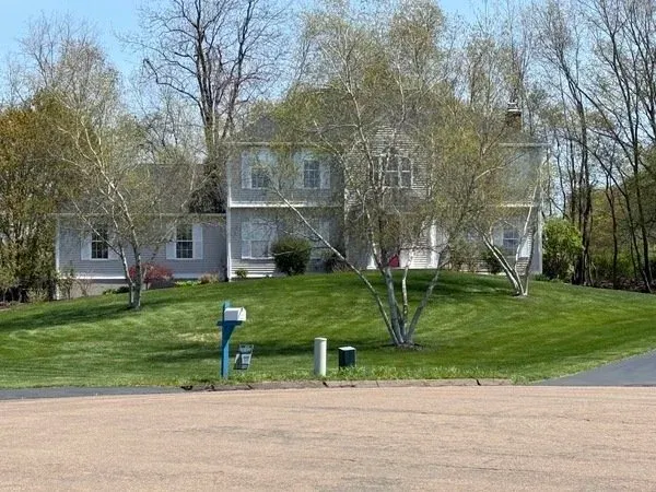 A white two-story house with multiple windows, surrounded by leafless trees and a well-maintained green lawn, at the corner of a street.