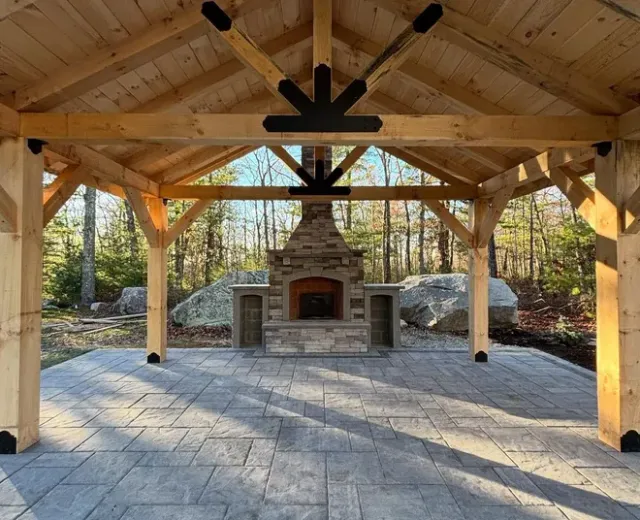 Wooden pavilion with a stone fireplace, on a gray stone patio. Forest in the background.