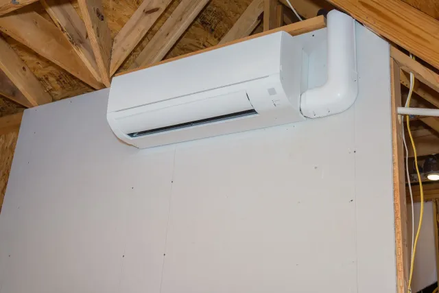 Wall-mounted white mini-split air conditioner on an unfinished interior wall with exposed wooden rafters.