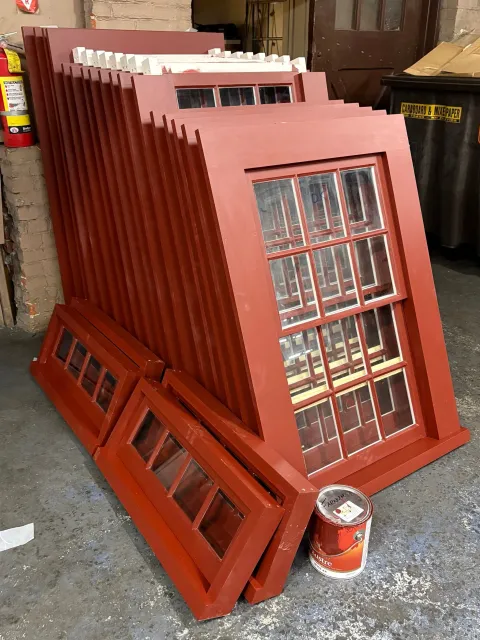 Stacked red-painted window frames with multiple glass panes leaning against a wall, next to a red paint can in a workshop or storage space.