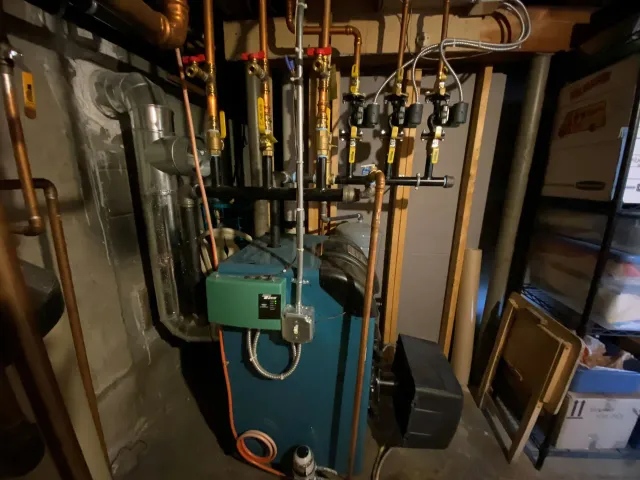 A residential basement in MA with a blue-green boiler system, various copper pipes, valves, and electrical wiring. Cardboard boxes and storage shelves are visible in the background—typical of Suffolk County or Norfolk homes.