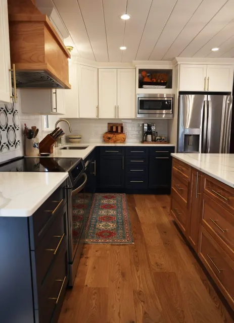 A modern kitchen with white upper cabinets, black lower cabinets, and a large wooden island. Features stainless steel appliances including a refrigerator, microwave, and oven, with a patterned backsplash and hardwood floor.