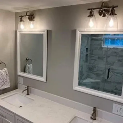 A modern bathroom with a double sink vanity, two rectangular mirrors, wall-mounted light fixtures above each mirror, gray walls, and a glass shower enclosure in the reflection.