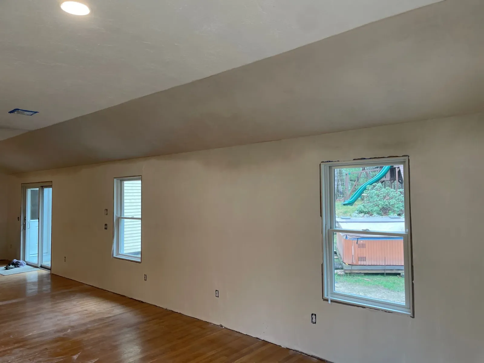 Interior of a room under renovation with beige walls, hardwood floors, and multiple windows, including a sliding door, leading outside to a backyard with a slide and hot tub.