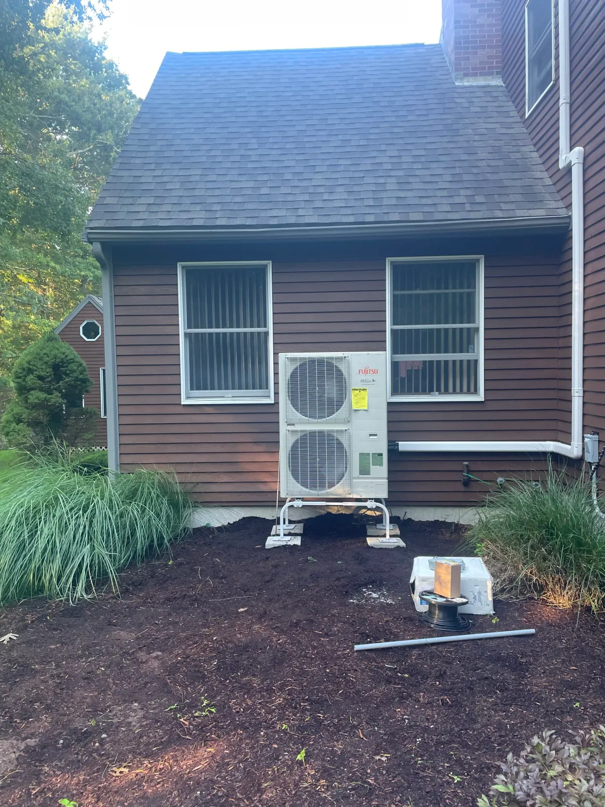Backyard view of a brown house with two windows, an outdoor air conditioning unit, and landscaped plants, with gardening tools and soil in the foreground.