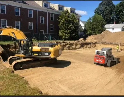 Construction site with a yellow excavator and a small red and white roller compactor on a dirt lot, surrounded by buildings and trees.
