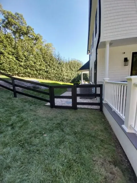 Black wooden fence near a white house and porch, with green grass and a gravel area.