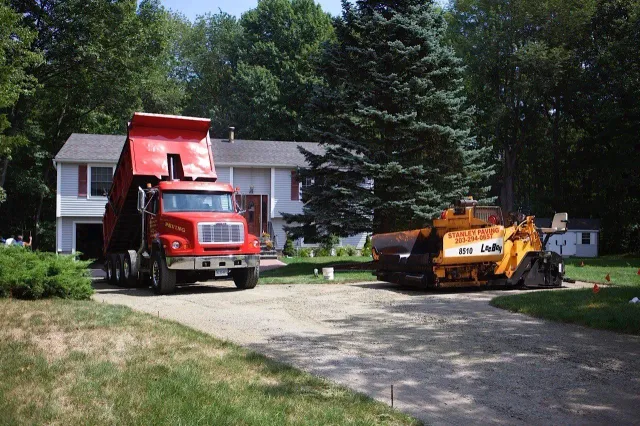 A red dump truck and yellow construction vehicle parked on a gravel driveway in front of a house.