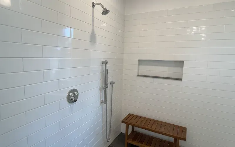 Minimalist shower with white subway tile walls, a rainfall showerhead, a handheld shower fixture, a recessed shelf, and a wooden bench.