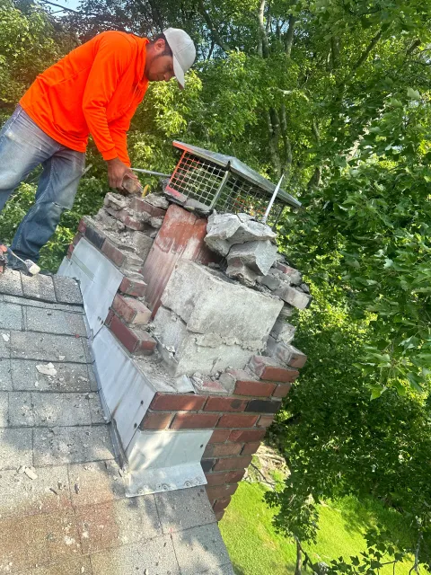 A worker in an orange long-sleeved shirt repairs a damaged brick chimney on a shingled roof surrounded by trees.