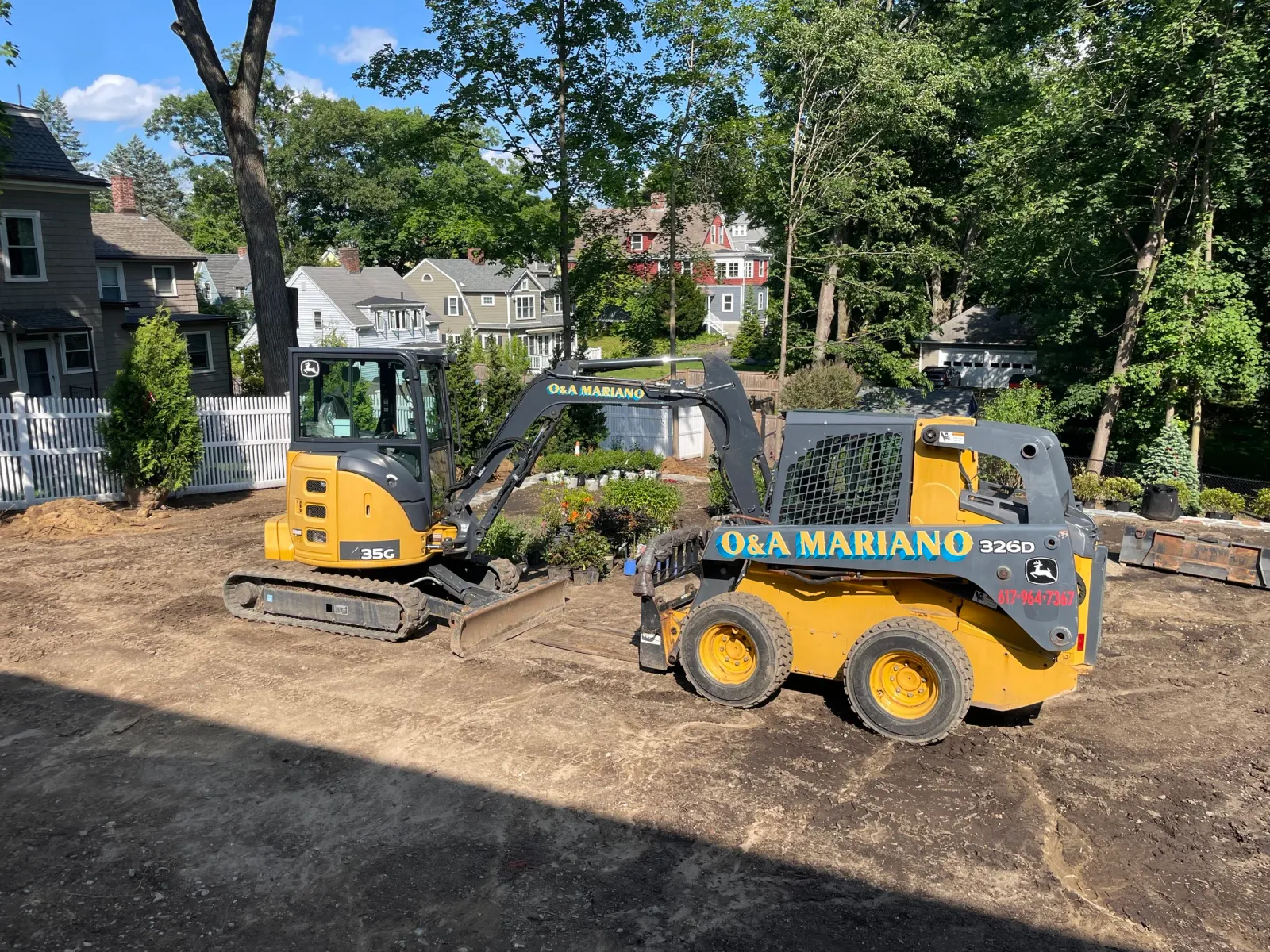 Construction site with a small yellow and black excavator and a compact loader with the company name 'Q&A MARIANO' on it, on dirt ground with residential houses and trees in the background.