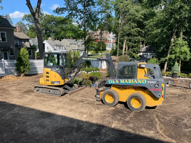 Construction site with a small yellow and black excavator and a compact loader with the company name 'Q&A MARIANO' on it, on dirt ground with residential houses and trees in the background.
