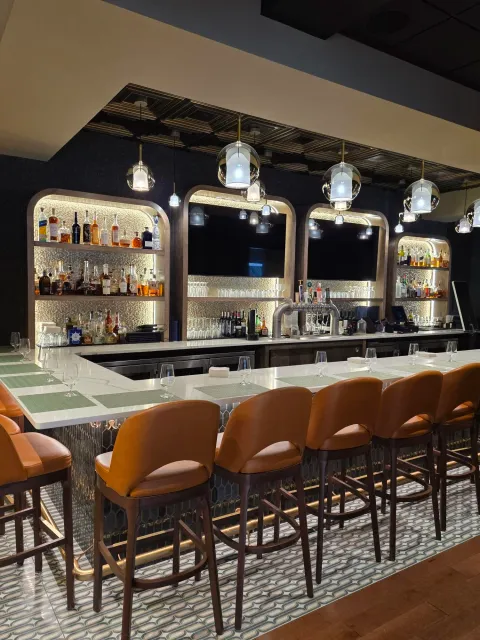 The image shows a modern bar with a white counter, brown bar stools, and a backlit shelf displaying liquor bottles. There are six hanging pendant lights with glass coverings above the bar, and a large flat-screen TV mounted on the wall behind the shelves. The flooring has a patterned tile design.