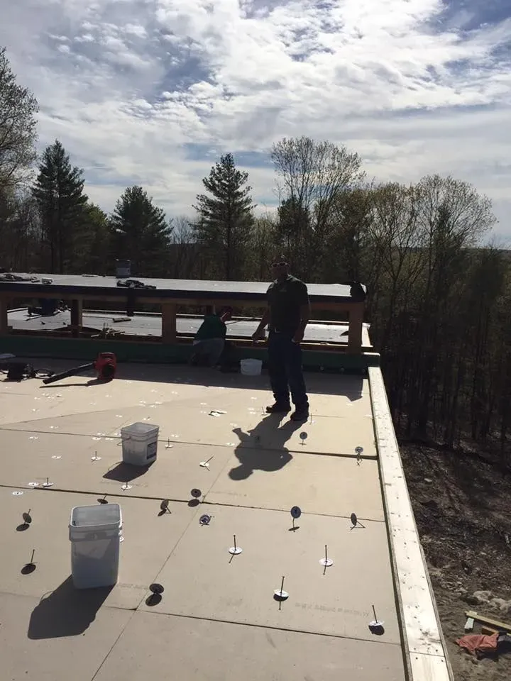 Two people on a flat roof under construction; one standing, one seated. Light colored sky with scattered clouds.