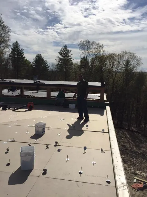 Two people on a flat roof under construction; one standing, one seated. Light colored sky with scattered clouds.