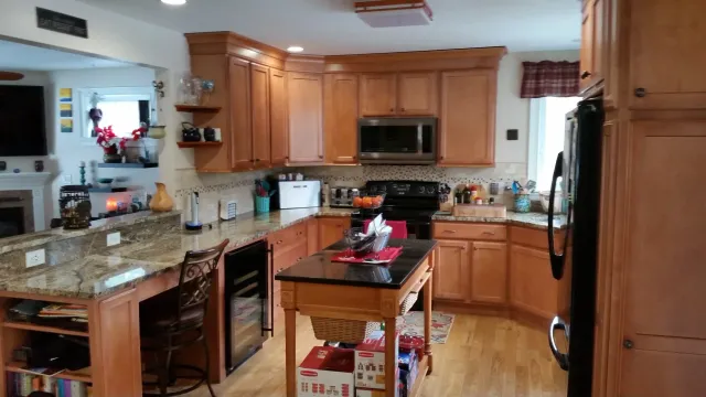 A kitchen with wooden cabinets, a granite countertop, a small island, a stove, microwave, and refrigerator. There is a window with a red checkered valance, various kitchen items, and a small cart with boxed items underneath.