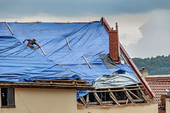 Damaged blue tarp roof with exposed beams and chimney after storm, over a house near water