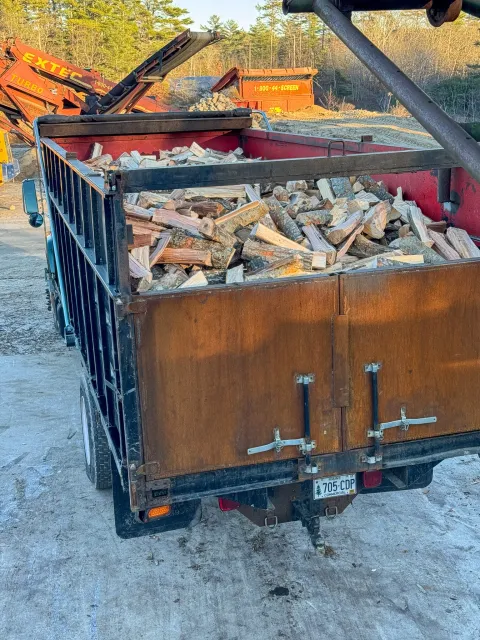 A trailer filled with chopped firewood at an outdoor lumber yard with other equipment and wood piles in the background. Tree Service Maine, Learned Tree Service