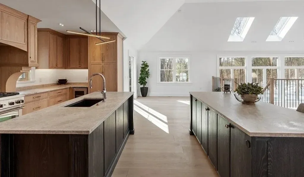 Open-concept kitchen with wooden cabinets, a large island with dark lower cabinets, and a light-colored countertop. The space is bright with natural light from large windows and skylights, and has a minimalist decor style.