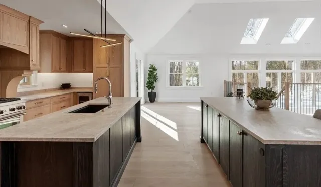 Open-concept kitchen with wooden cabinets, a large island with dark lower cabinets, and a light-colored countertop. The space is bright with natural light from large windows and skylights, and has a minimalist decor style.