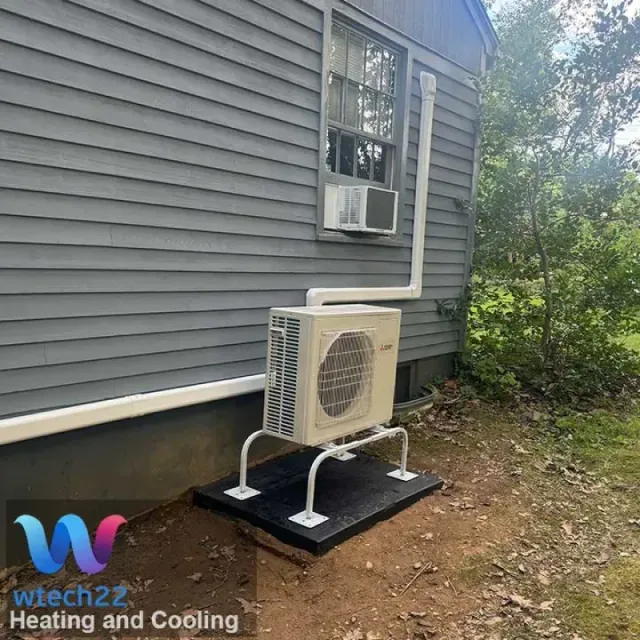 An outdoor HVAC unit on a platform, attached to a house with white piping and an above window unit.