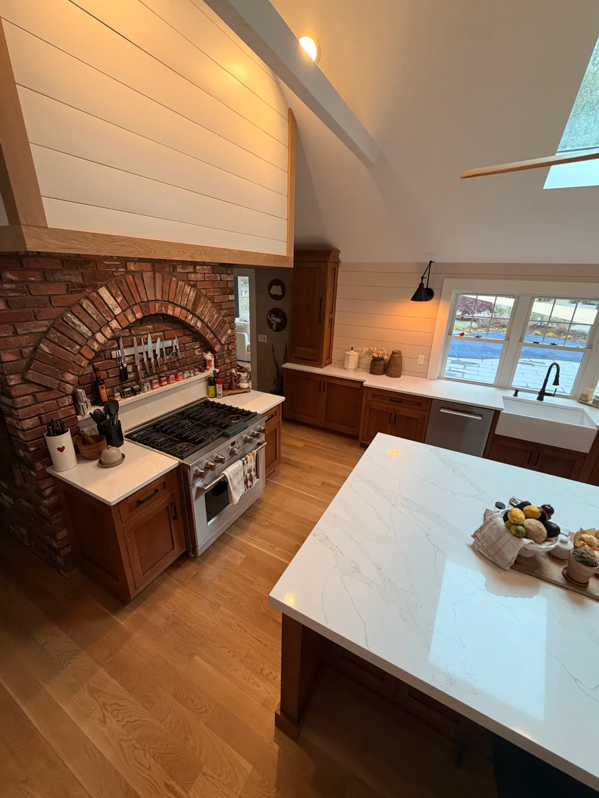 A modern kitchen with a white marble countertop, wooden cabinets, a brick arch over the stove area, a window with a view outside, and various kitchen utensils and decorative items.