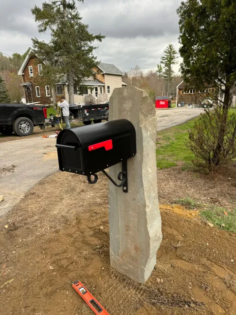 Black mailbox mounted on a concrete post beside a dirt driveway in a suburban neighborhood.