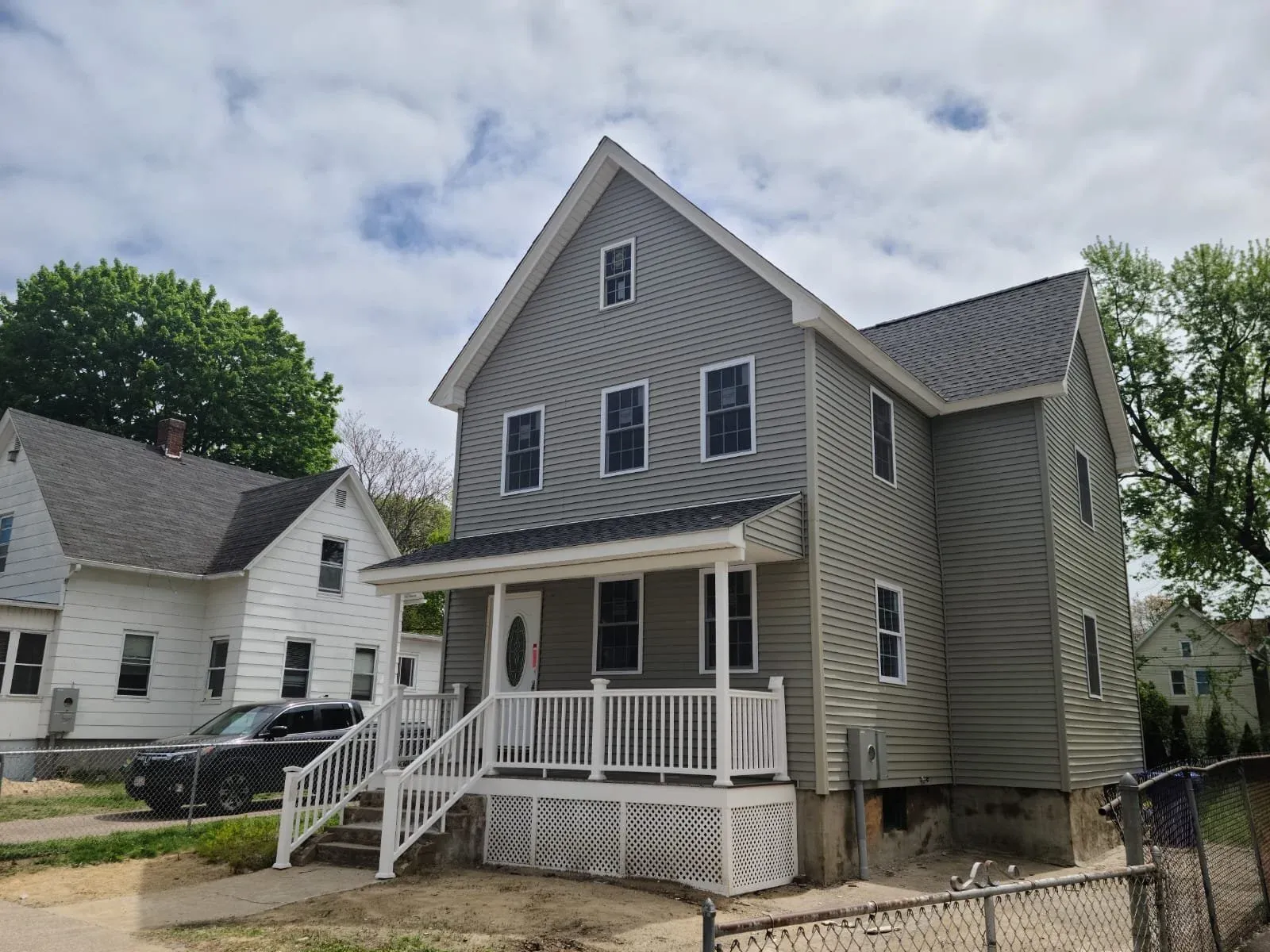 A new gray, three-story house with white trim, small front porch with railing and steps, and a driveway with a black vehicle parked on it. There are neighboring houses and trees, with some clouds in the sky.