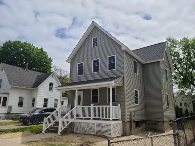 A new gray, three-story house with white trim, small front porch with railing and steps, and a driveway with a black vehicle parked on it. There are neighboring houses and trees, with some clouds in the sky.
