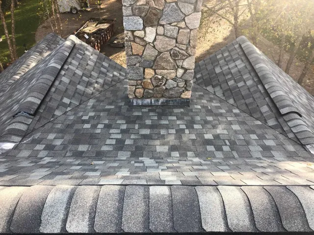 A rooftop with grey asphalt shingles and a prominent stone chimney in the center. The shingles are laid out in a neat, patterned arrangement. Sunlight partially lights the scene, casting shadows and highlighting the texture of the roofing materials.