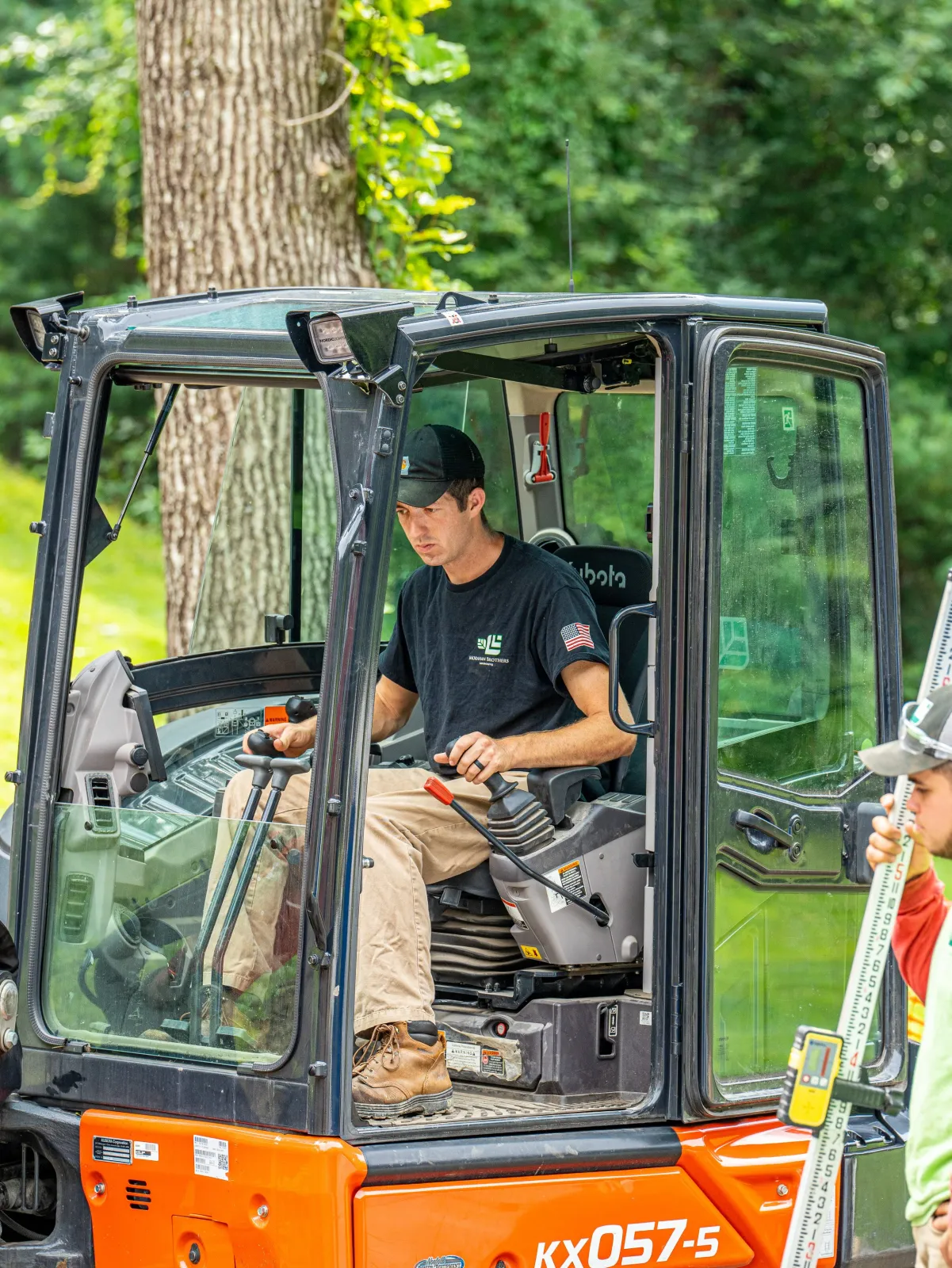 A man operating a small orange and black construction vehicle outdoors, with a woman holding a measuring stick nearby.