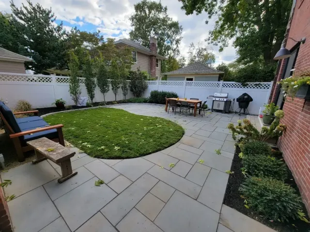 A tiered stone patio with outdoor furniture, landscaping, and a car parked on an upper level, seen from a paved driveway.