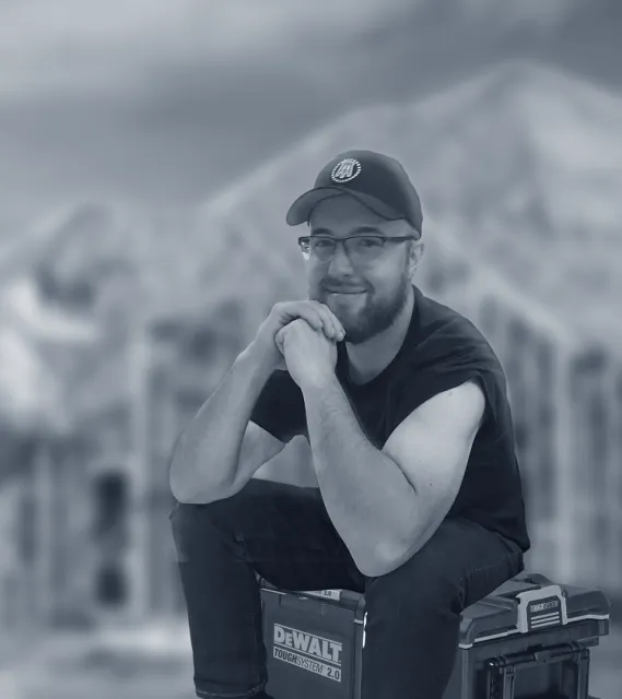 A cape cod electrician sitting on a toolbox looking kindly at camera. The background is an our of focus blurred house.