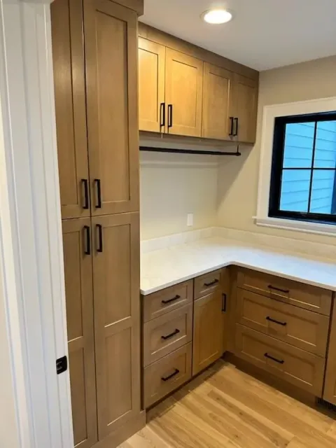 A custom-built wood pantry cabinet beside a matching counter with drawers and upper cabinets, under a small window.