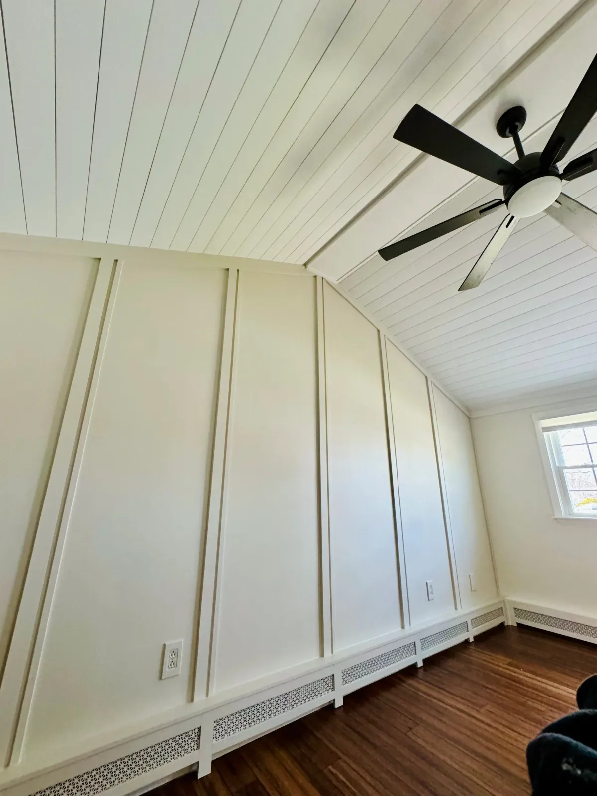 A room with white paneled walls and ceiling, a black ceiling fan, a window on the right wall, hardwood flooring, and a white baseboard heater along the bottom of the wall.