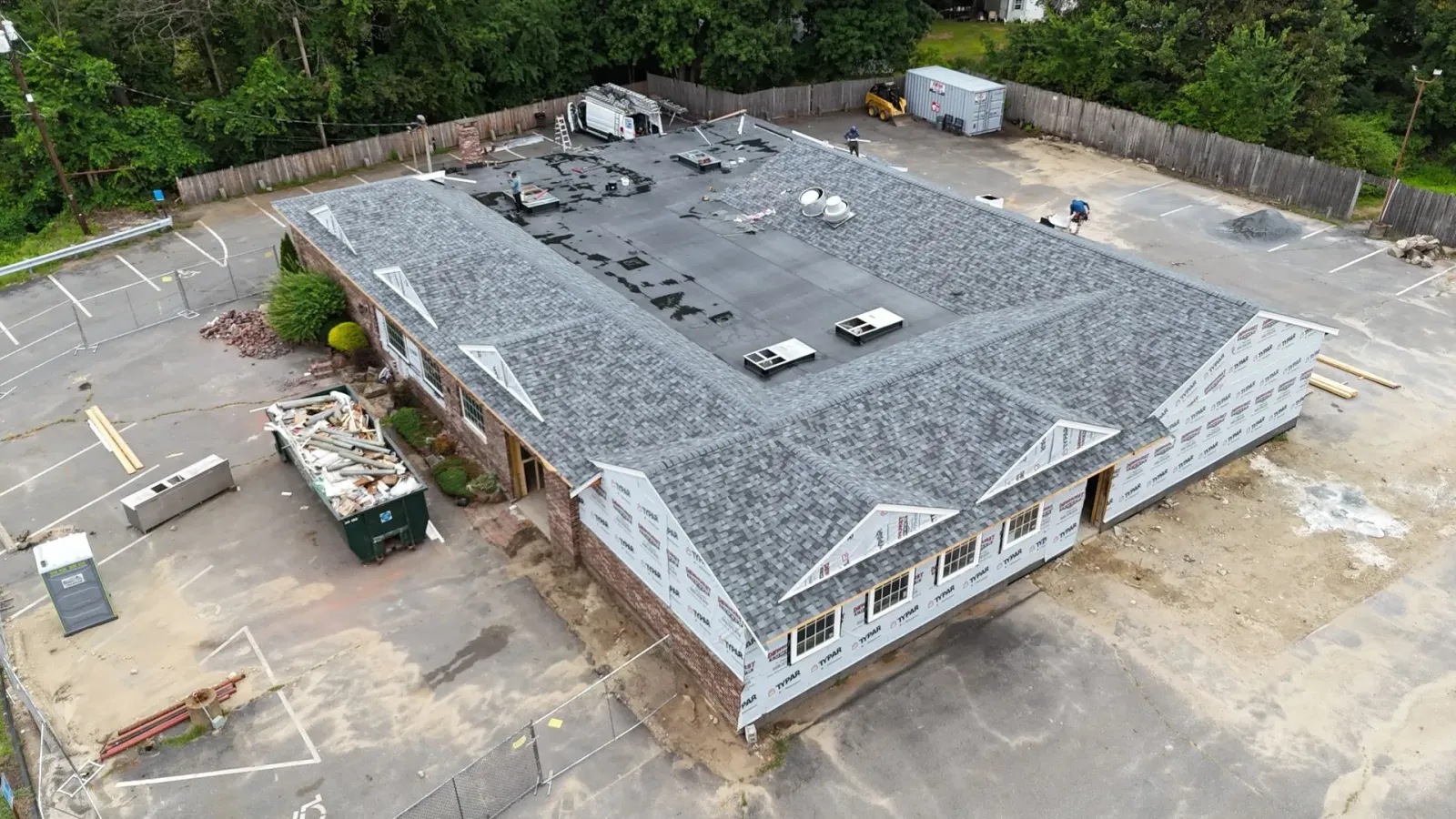 An aerial view of a rectangular building under construction, showing a new gray shingled roof and exterior wall wrap.