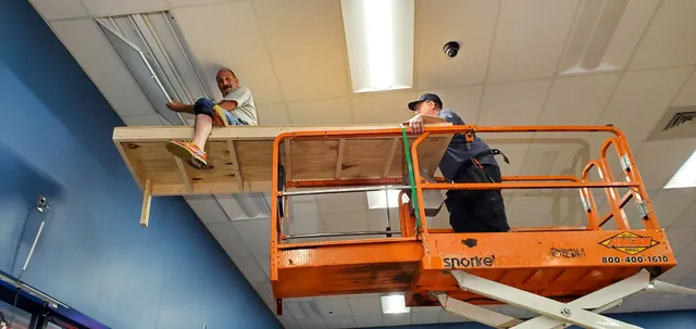 Two workers on an orange lift platform collaborate to install or repair ceiling fixtures in a commercial space.