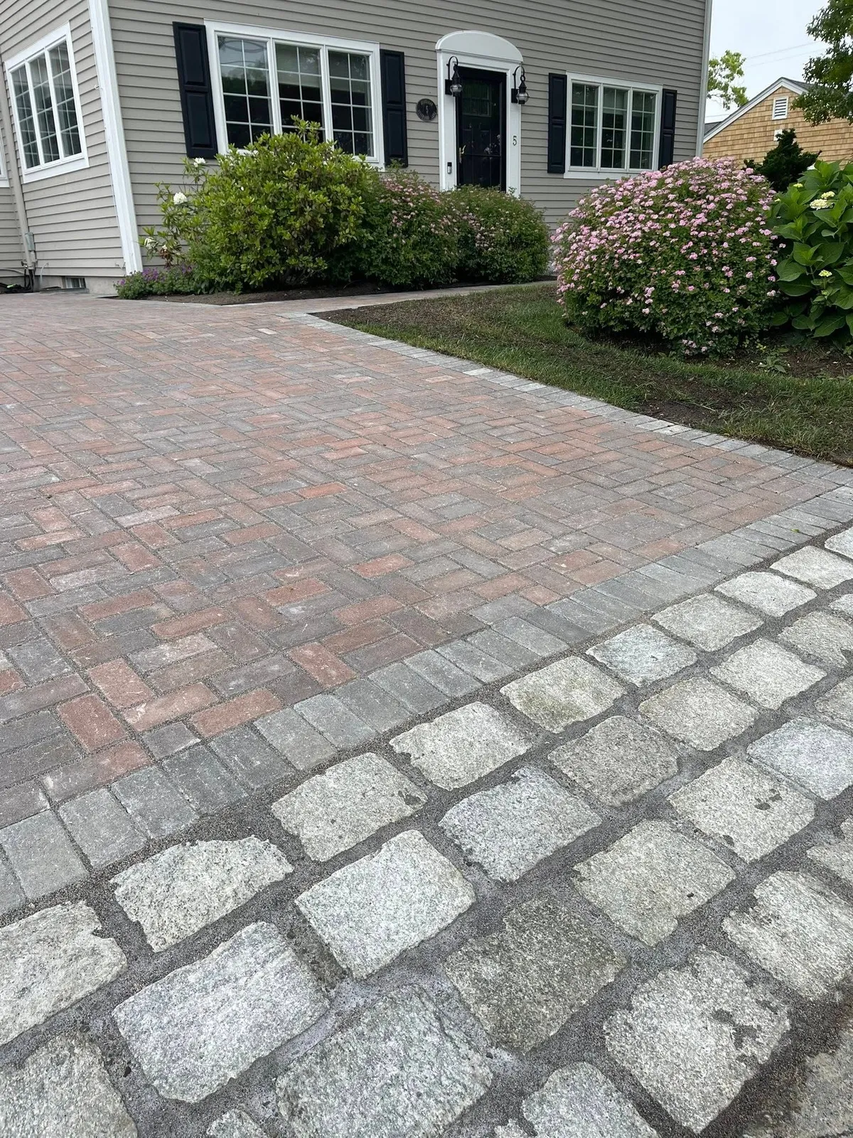 Brick paver driveway leading to a house with black shutters and door, edged with stone pavers. Green bushes flank the walkway.