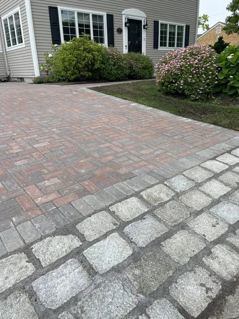 Brick paver driveway leading to a house with black shutters and door, edged with stone pavers. Green bushes flank the walkway.