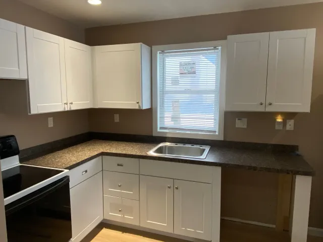 A kitchen with white cabinets , a stove , a sink and a window.