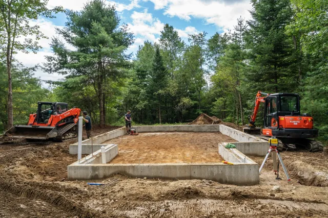 Construction site with concrete foundation and equipment, surrounded by trees, workers, and machinery including a small excavator and a roller compactor, under a partly cloudy sky.