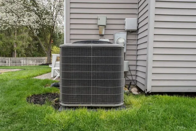 Air conditioning unit outside a house with gray siding, sitting on green grass.