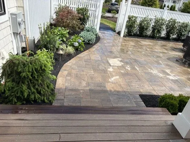 Stone patio with dark border, bordered by a garden path and stairs, leading to a white fence.