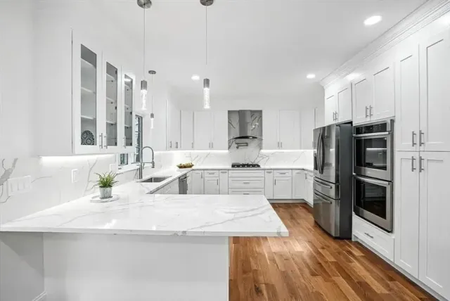 Modern white kitchen with marble countertops, stainless steel appliances, and hardwood floors.