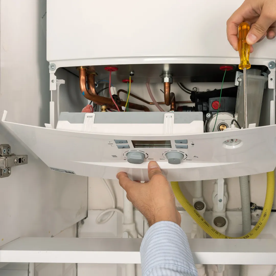 A person using a screwdriver to open the front panel of a boiler or water heater, exposing internal components like pipes and wires.