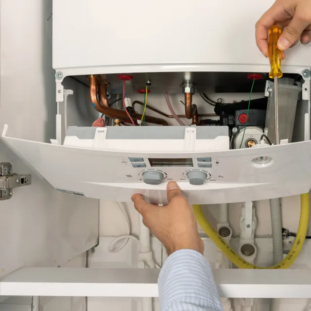 A person using a screwdriver to open the front panel of a boiler or water heater, exposing internal components like pipes and wires.